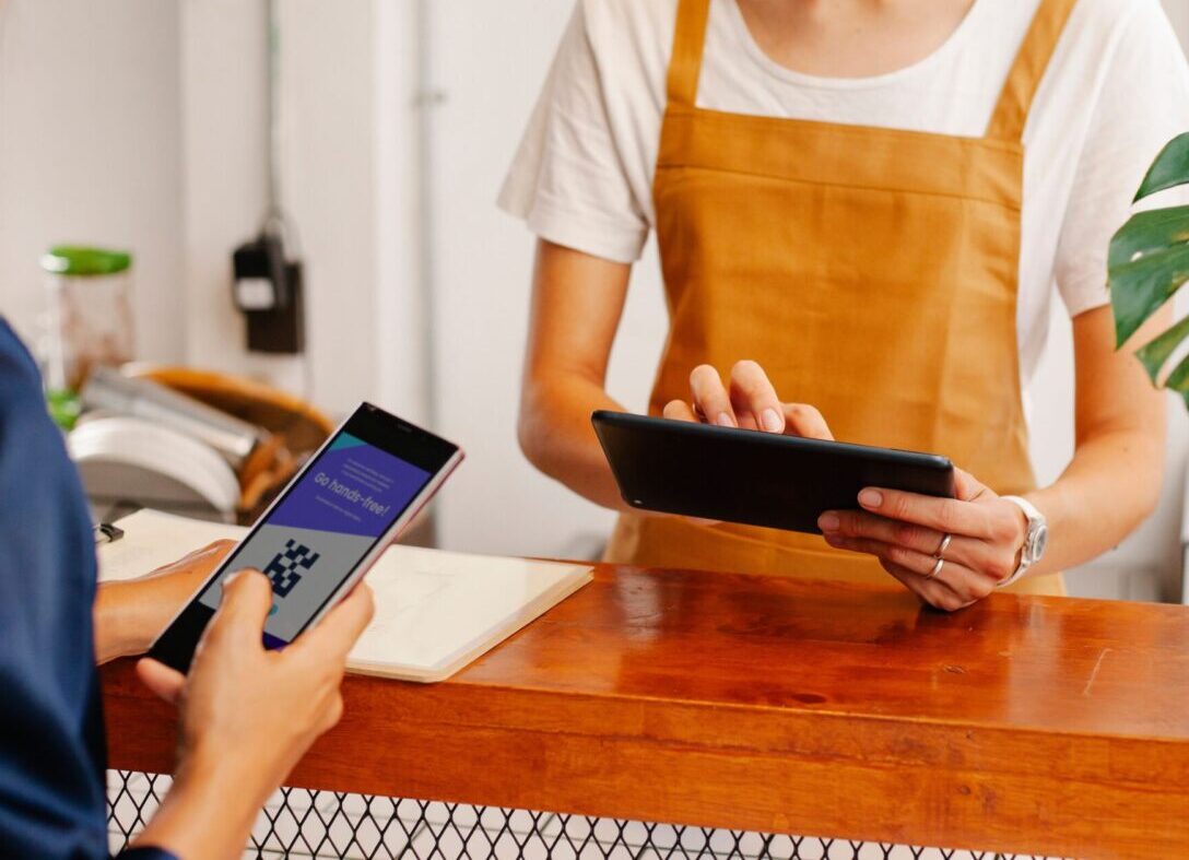Crop anonymous female employee with application on cellphone screen interacting with partner using tablet at counter in cafeteria